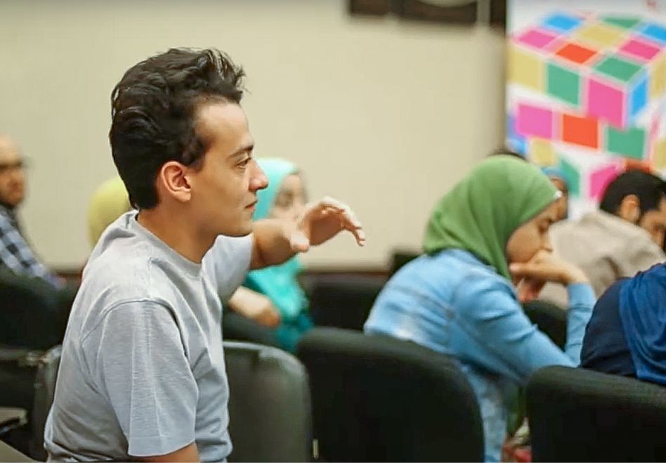 Photo: A young man raises his hand in a classroom. Other students are visible in the background, with the women wearing headscarves.