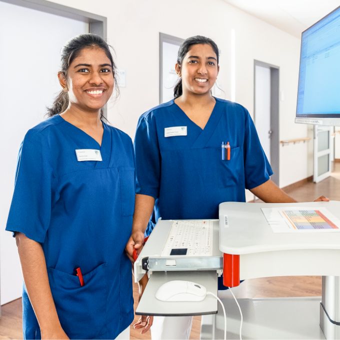 Photo: Two young women in dark blue scrubs standing in a hospital corridor next to a monitor.