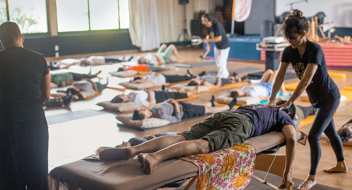 Photo: A man lies on his stomach on a massage table and is given a shoulder massage. In the background, people are lying on yoga mats.