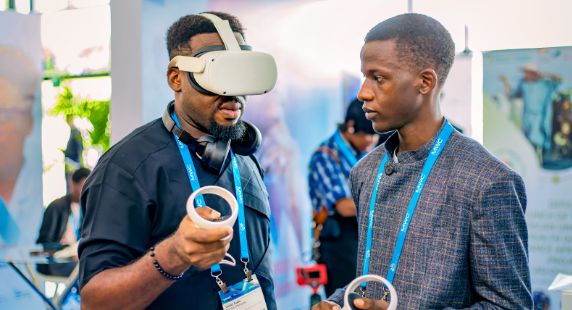 Photo: Two men standing face-to-face. The man on the left is wearing a VR headset.