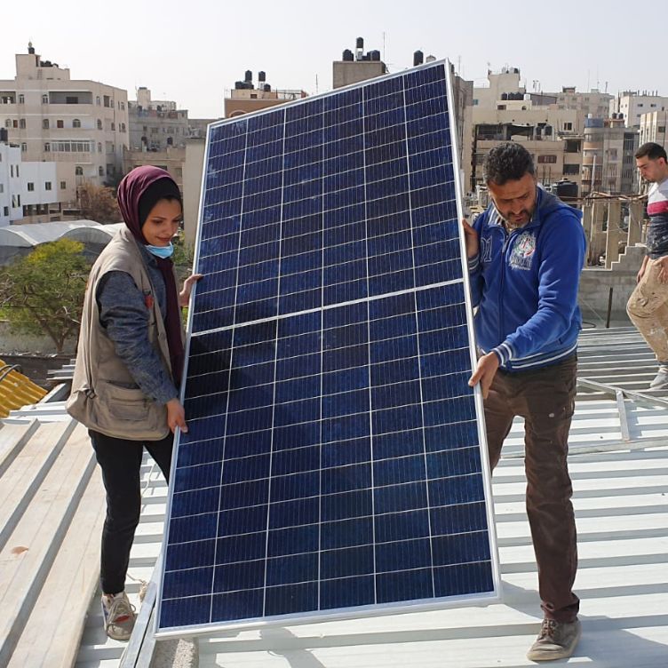 Photo: A man and a woman carrying a large solar panel across a roof.