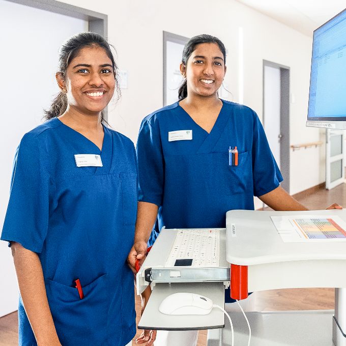 Photo: Two young women in dark blue scrubs standing in a hospital corridor next to a monitor.