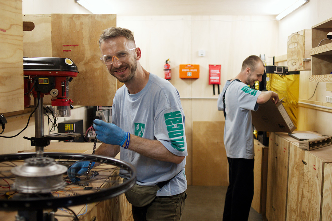 Photo: A man wearing protective goggles repairs a bicycle chain in a workshop. Another man is holding a folder. Both are smiling. &copy; GIZ / Sergiy Bezborodko