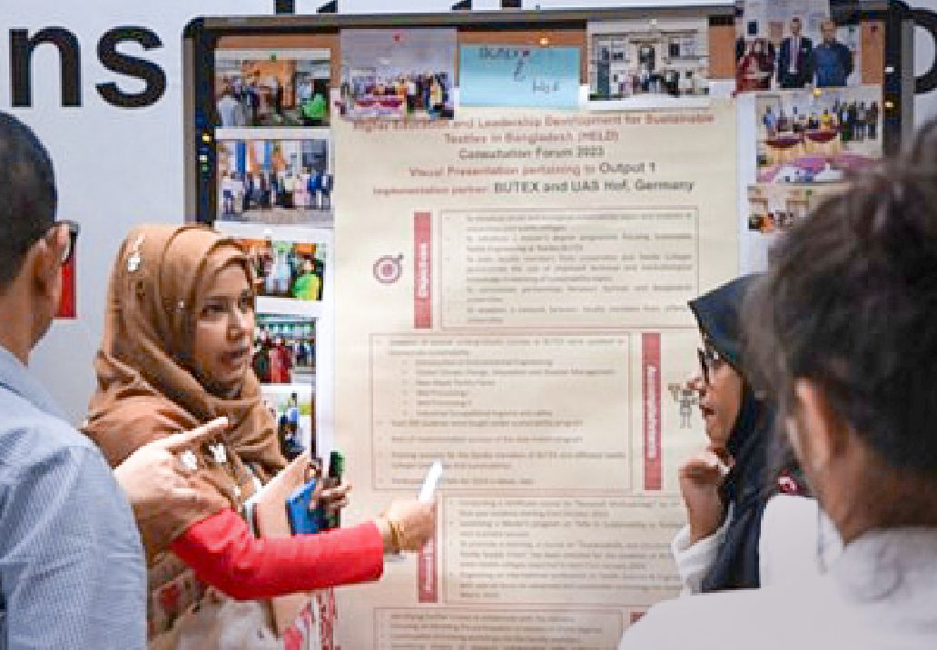 Photo: Four people standing in front of a notice board. A woman points to it and speaks.