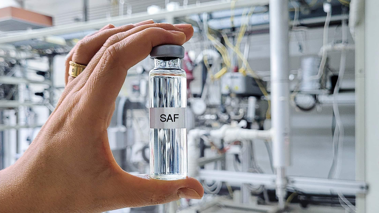 A hand holding up to the camera a sealed test tube labelled ‘SAF’. In the background, there are many interconnected pipes and cables.