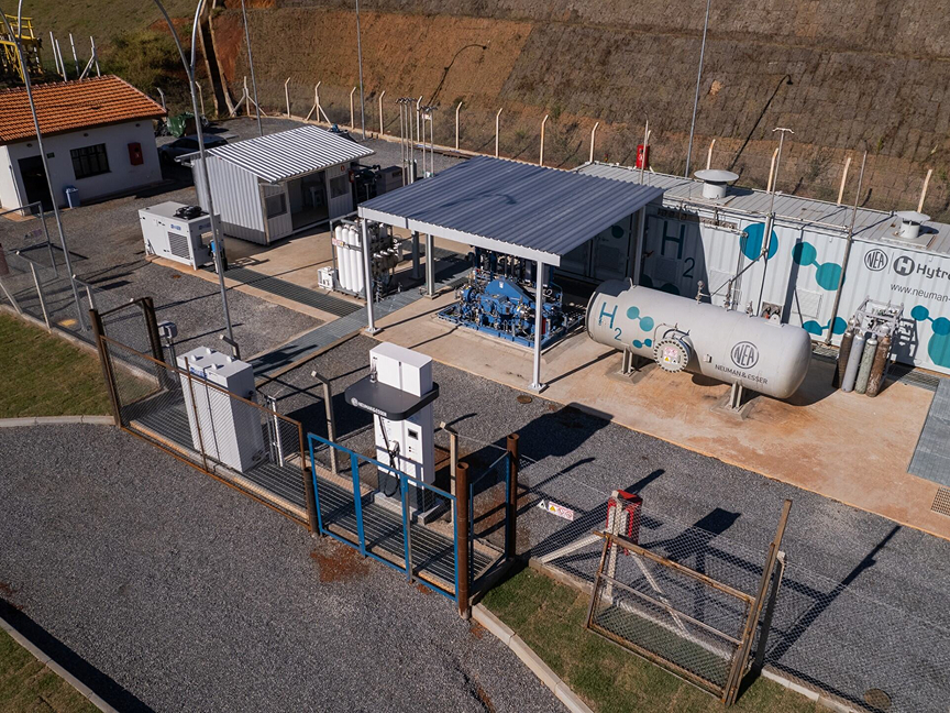 A fenced-in pilot plant for the production of green hydrogen located on the edge of a hillside. ‘H2’ is written on one of the various interconnected storage tanks.