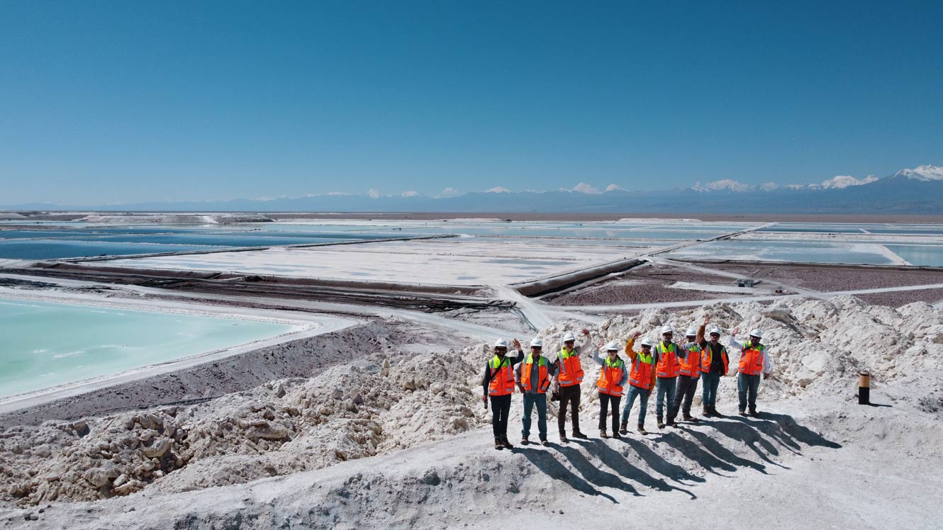 A group of people in protective clothing standing in front of a vast salt flat with evaporation ponds in a mountainous area.