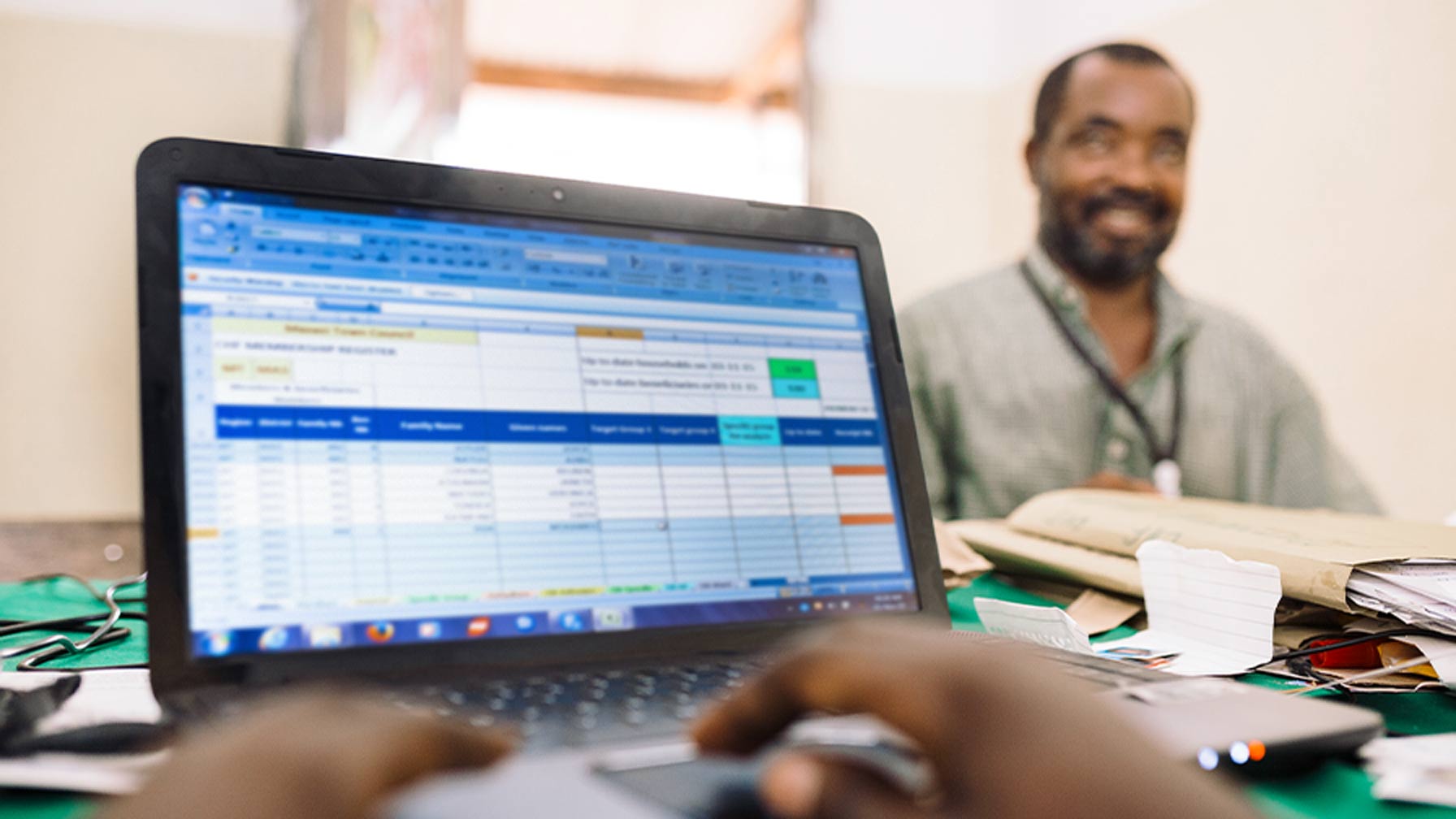 A person typing on a laptop showing an Excel table; in the background, a man sits at a desk smiling.
