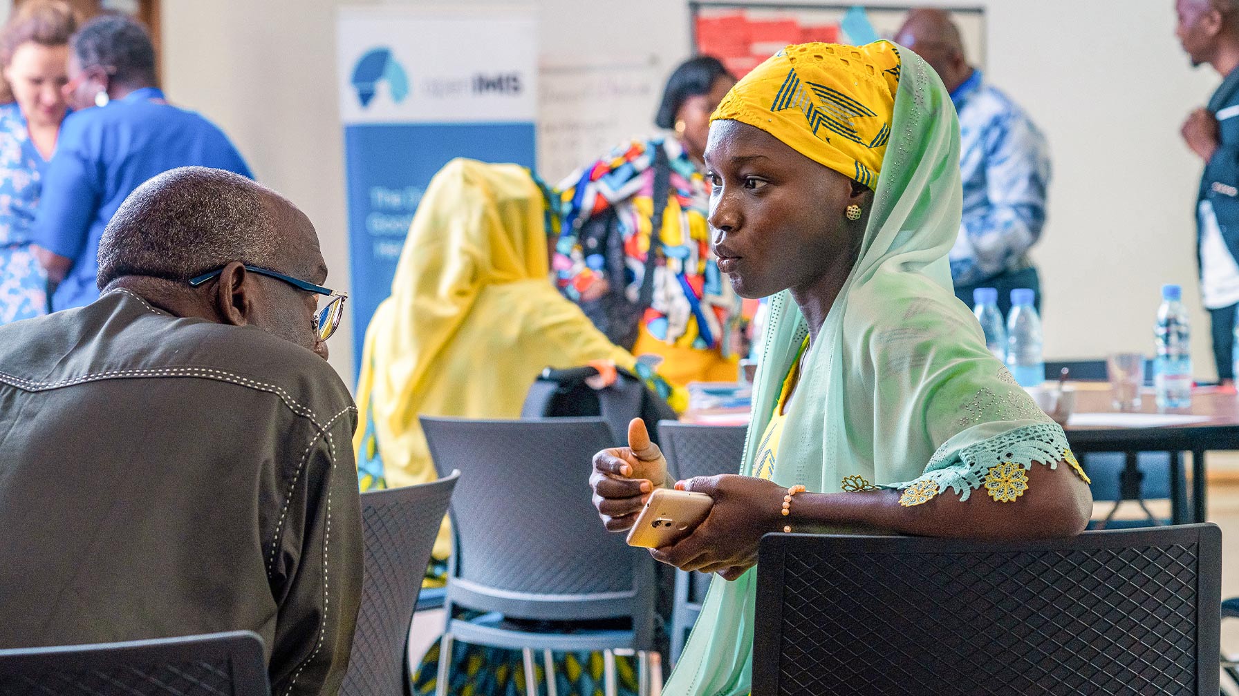 A woman in brightly coloured clothing talks to a man at an event in Cameroon; other participants can be seen in the background.
