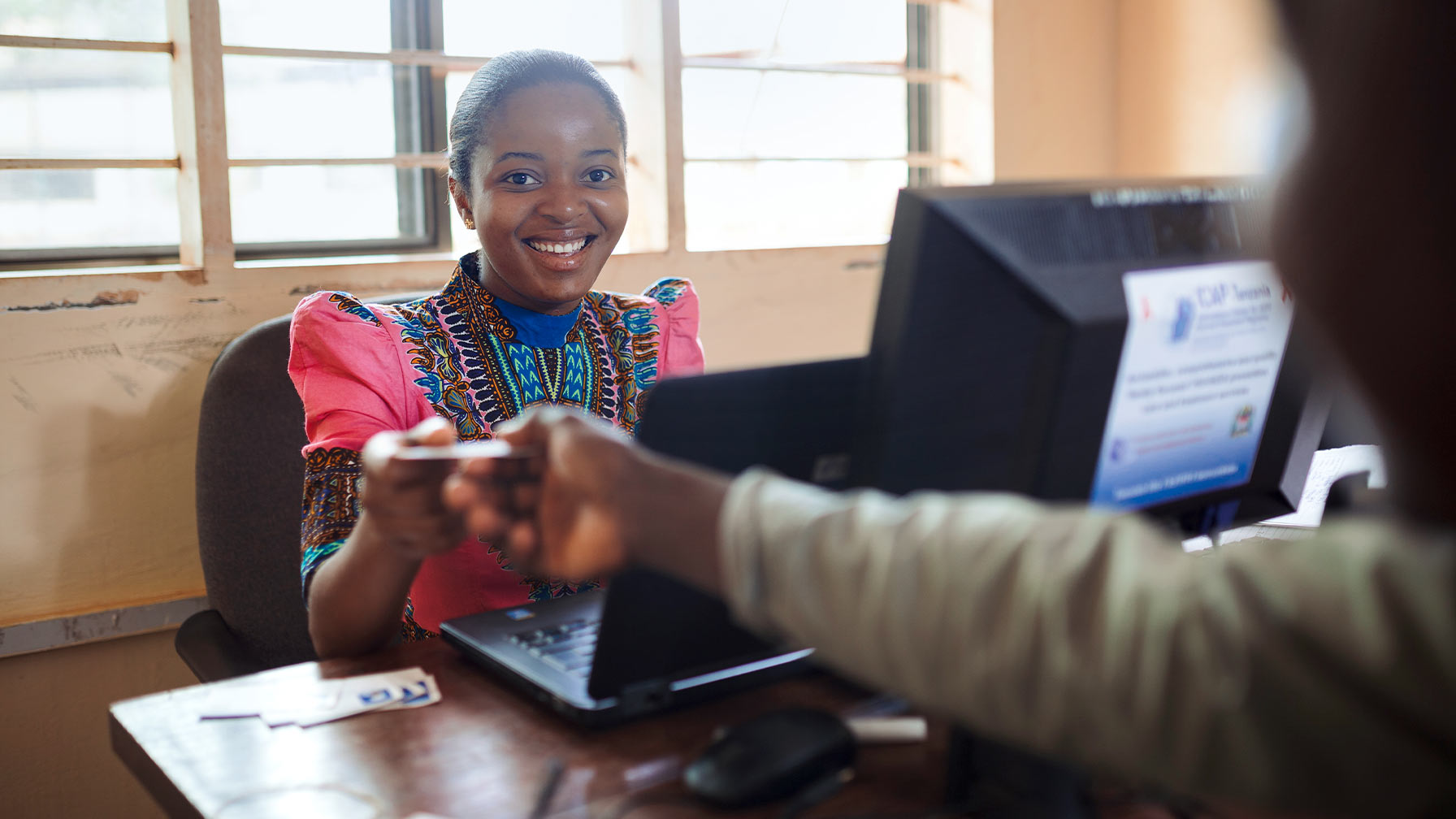 A smiling woman sitting in an office in Tanzania hands over a card to someone.