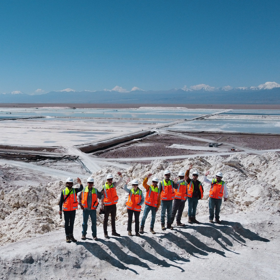 A group of people in protective clothing standing in front of a vast salt flat with evaporation ponds in a mountainous area.