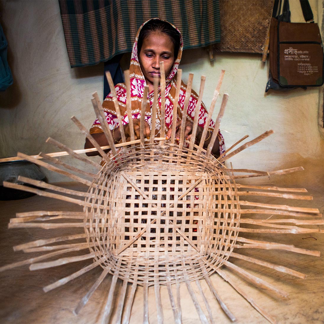 A woman sits in a room and weaves a large basket from wooden strips with her hands.