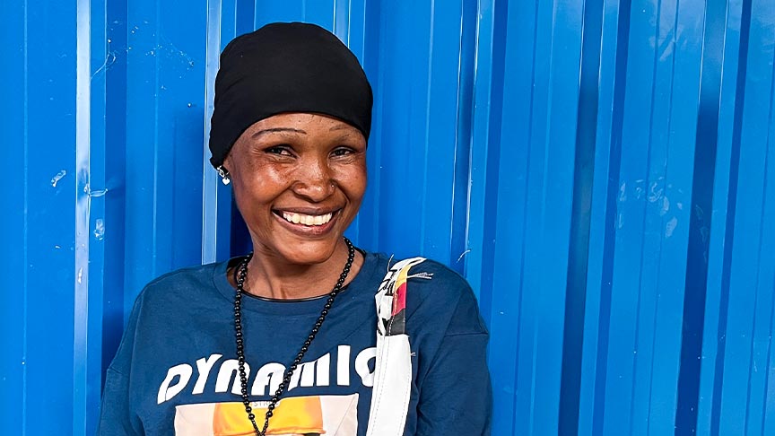 A smiling woman stands in front of a blue metal wall wearing a printed t-shirt and carrying a brightly coloured fabric bag with a floral pattern on it.