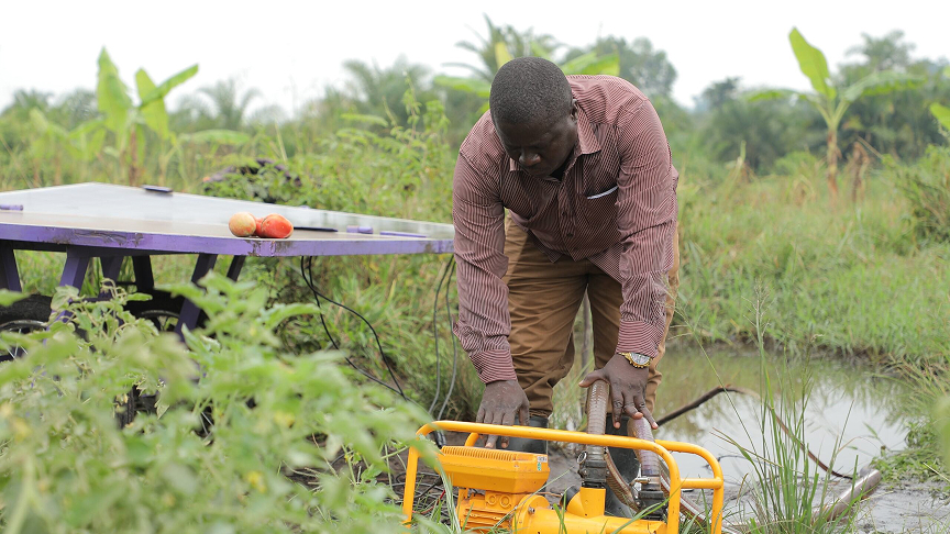 A man working in a field at a yellow water pump; next to it is a table with a solar panel and two tomatoes on it.