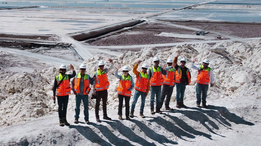 A group of people in protective clothing standing in front of a vast salt flat with evaporation ponds in a mountainous area.