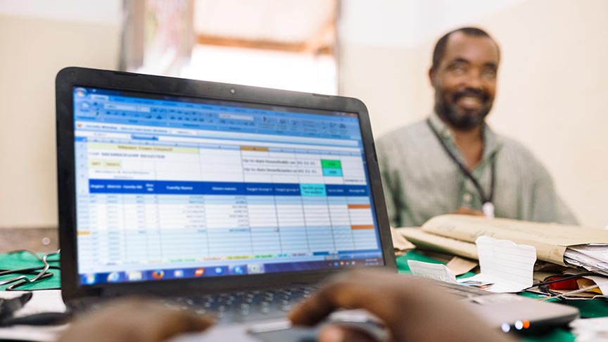 A person typing on a laptop showing an Excel table; in the background, a man sits at a desk smiling.