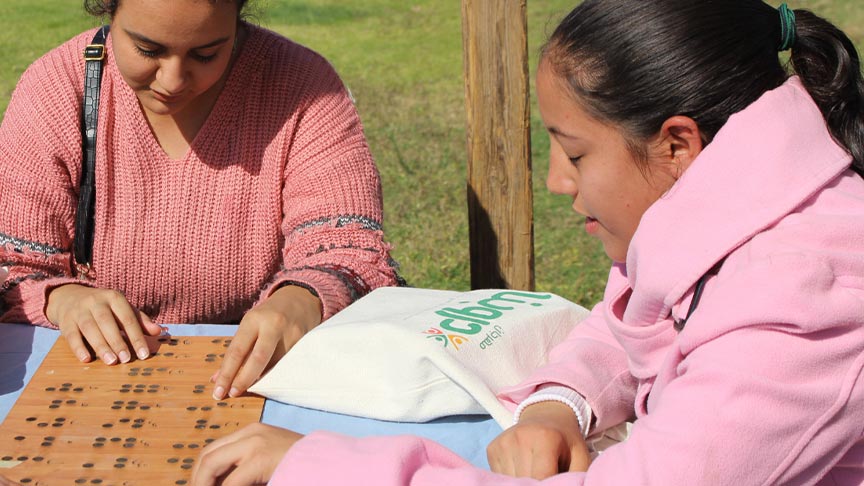 Three young women sitting at a table outdoors run their fingers over a wooden board with Braille.