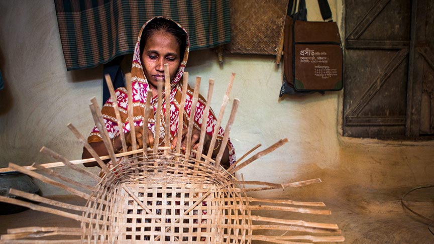 A woman sitting in a room hand-weaves a large basket made of wooden strips.