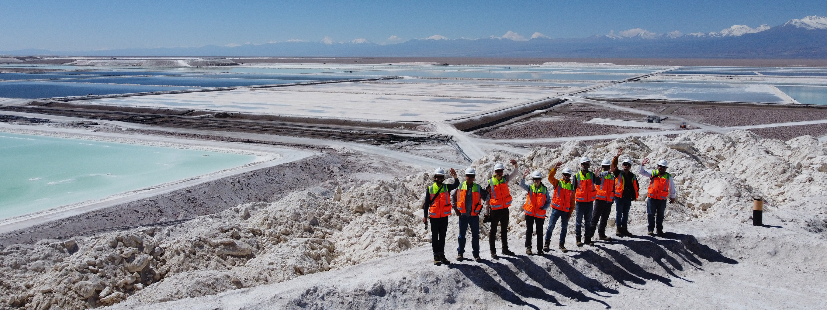 A group of people in protective clothing standing in front of a vast salt flat with evaporation ponds in a mountainous area.