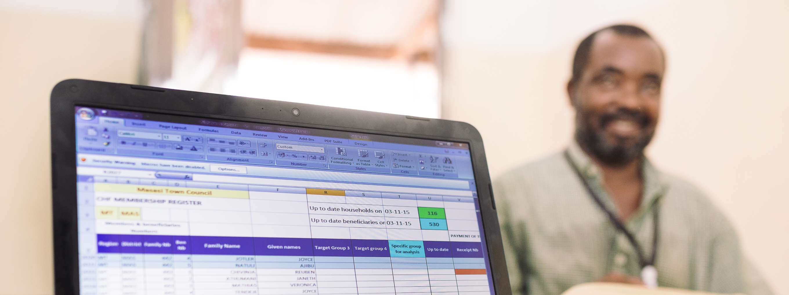 A person typing on a laptop showing an Excel table; in the background, a man sits at a desk smiling.