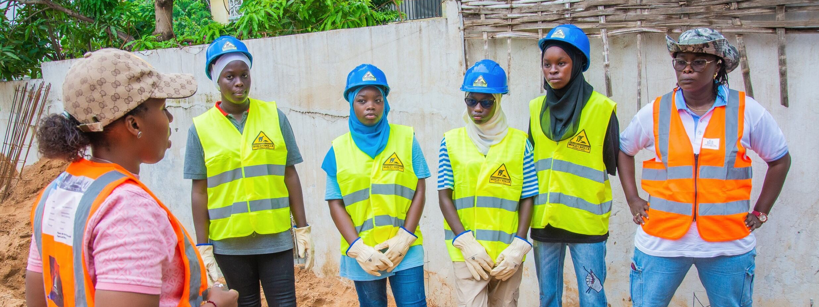 Group photo with Djélia Diagne (in profile on the left) talking to five young women in hard hats and high-visibility vests on a construction site.