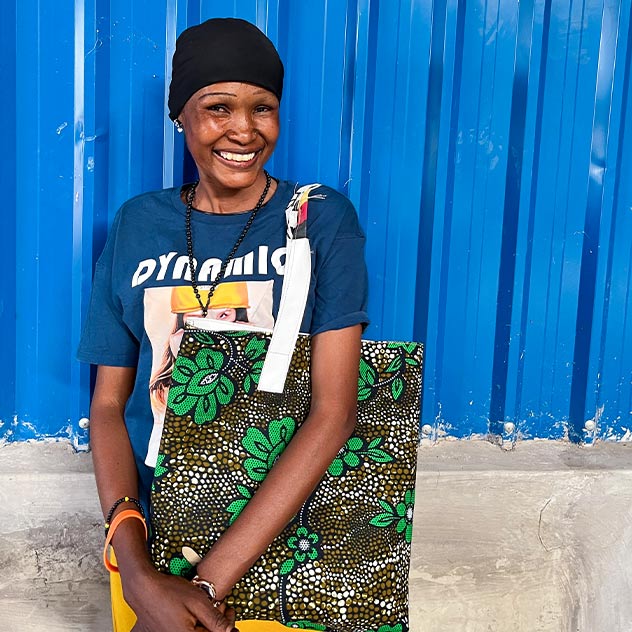 A smiling woman stands in front of a blue metal wall wearing a printed t-shirt and carrying a brightly coloured fabric bag with a floral pattern on it.