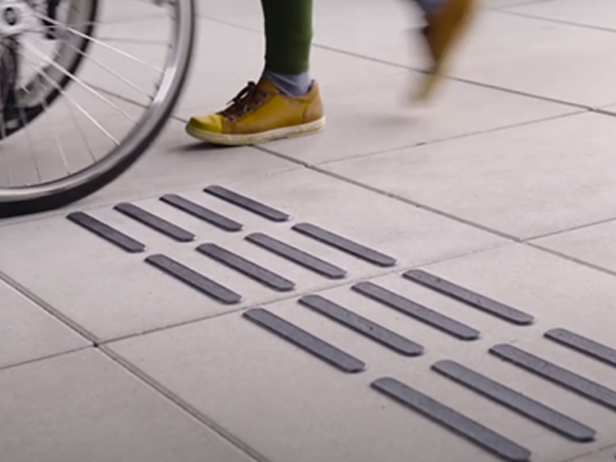 Close-up of a walkway with tactile guiding strips for people with visual impairments, with a wheelchair and a person walking above it.