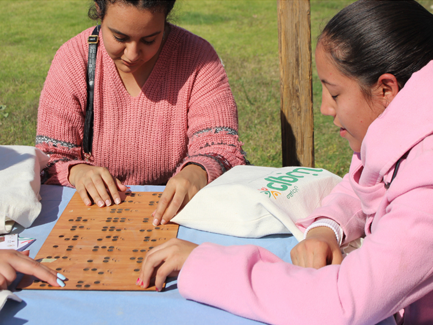 Three young women sitting at a table outdoors run their fingers over a wooden board with Braille.