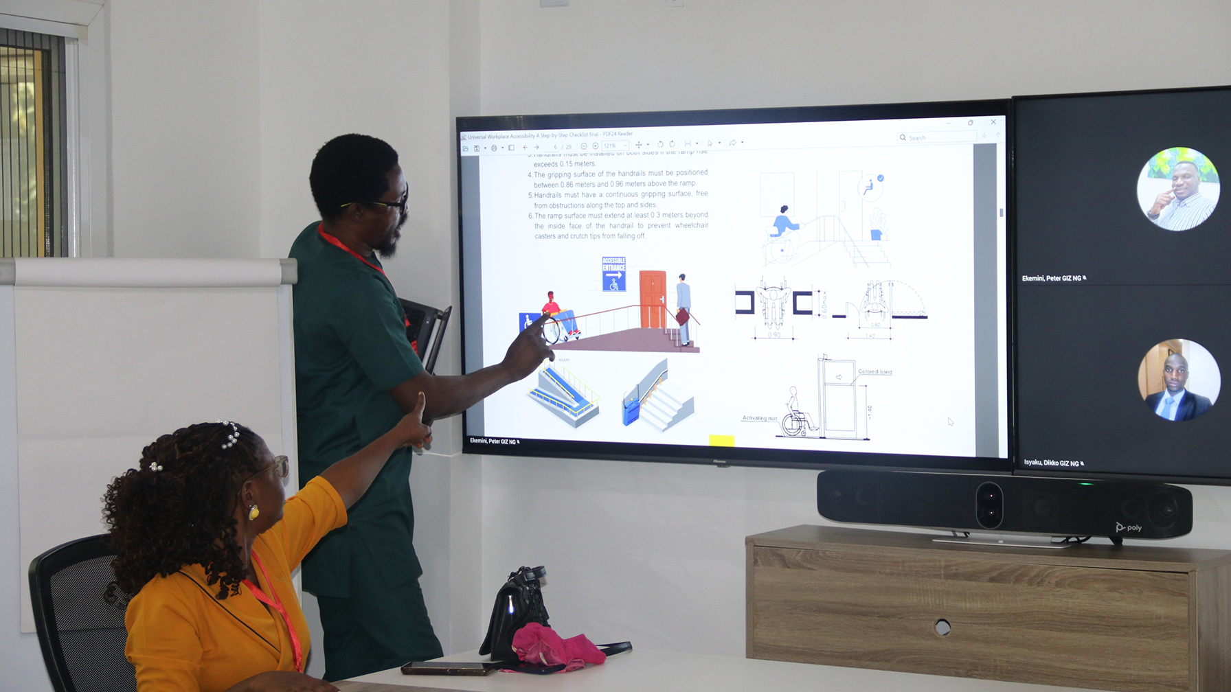 A man and a woman discussing a diagram on accessible infrastructure at a screen while two other participants join the meeting virtually.
