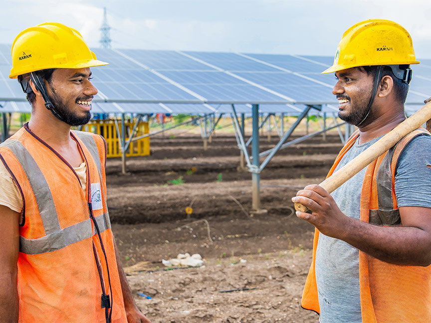 Two men in hard hats and high-visibility vests stand smiling on a solar field; one is holding a spade over his shoulder.
