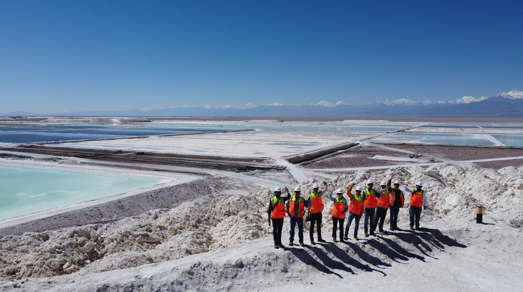 A group of people in protective clothing standing in front of a vast salt flat with evaporation ponds in a mountainous area.