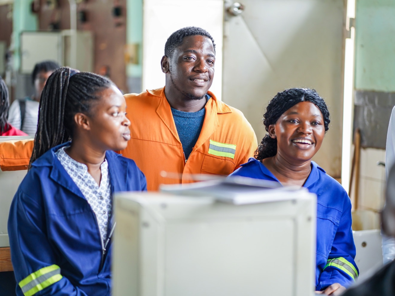 Three people in overalls standing or sitting in a workshop or training room. They are smiling and surrounded by other people.