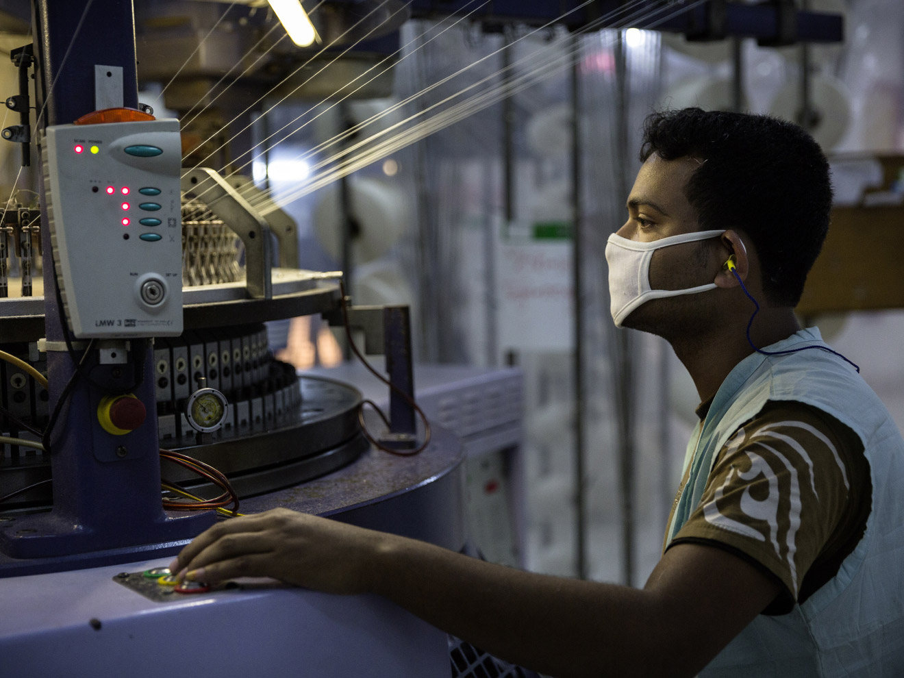 A man wearing a mask and hearing protection operates an industrial textile machine, keeping a close eye on the production process.