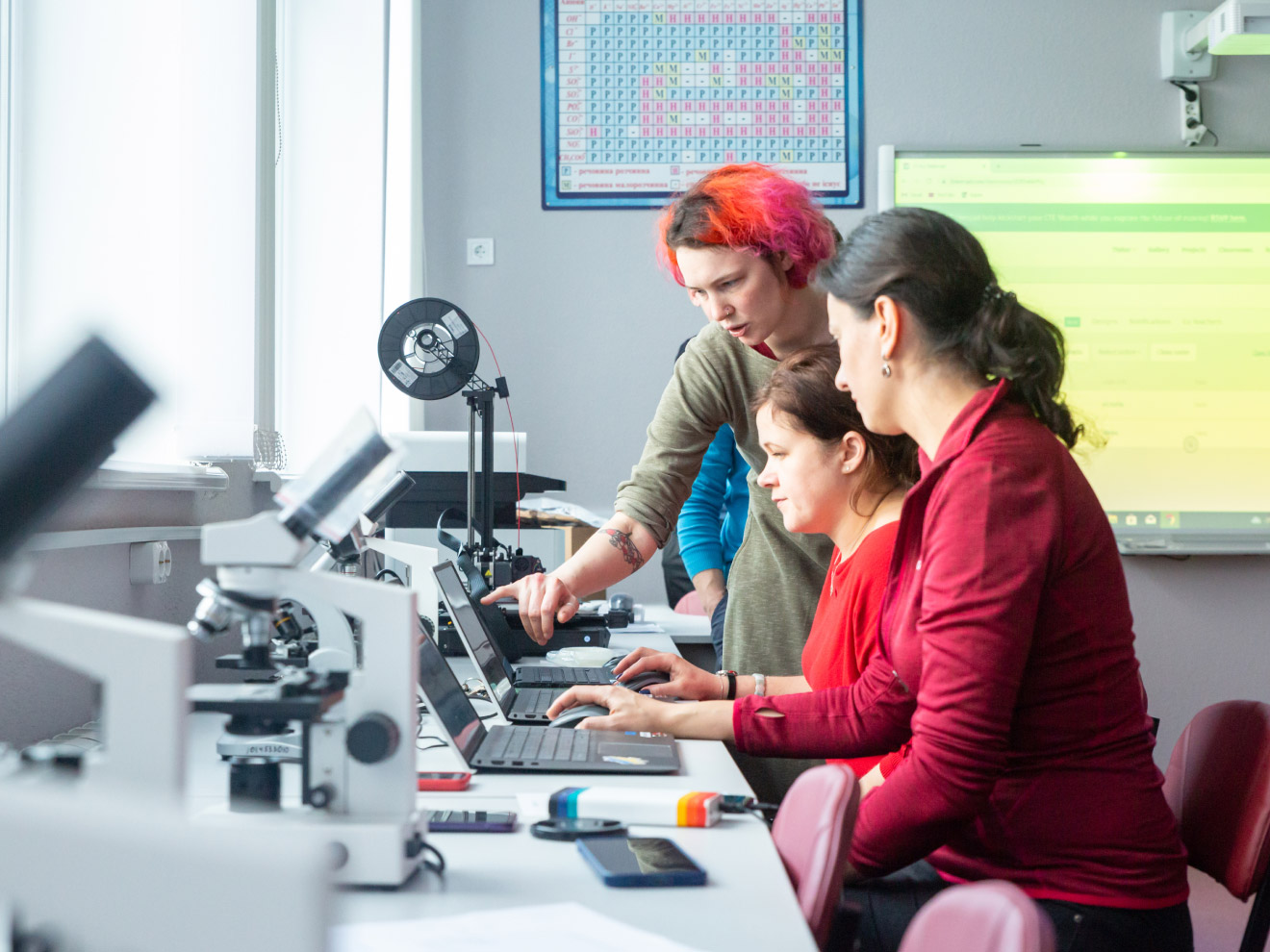 Three people working on laptops in a classroom with microscopes; a fourth person is explaining something.