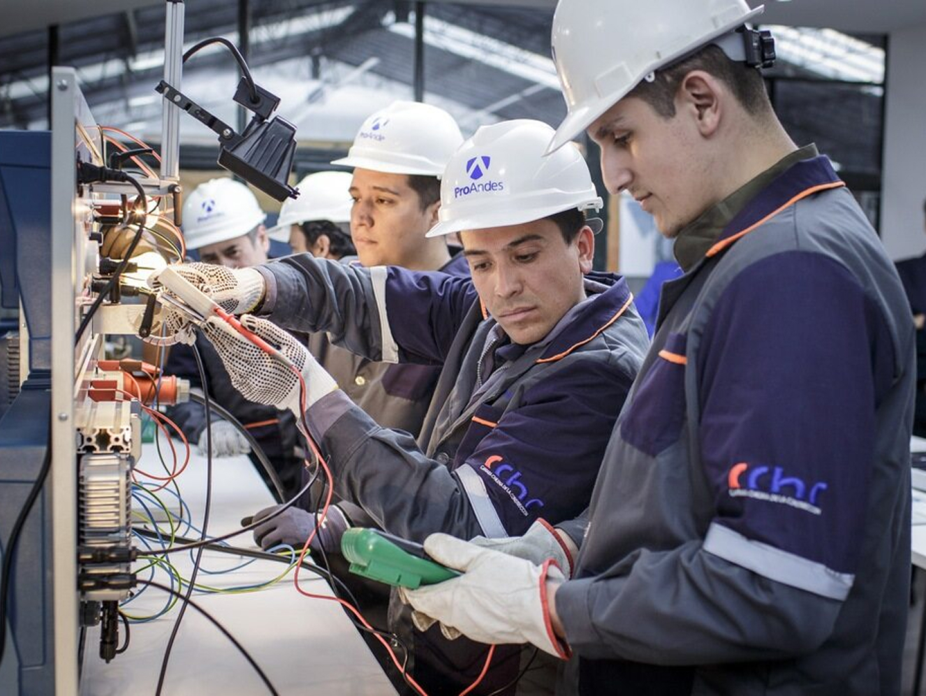 Several men in protective clothing and hard hats working intently on an electric circuit in a workshop.